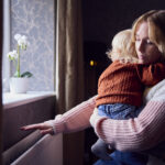 Mother With Son Trying To Keep Warm By Radiator At Home